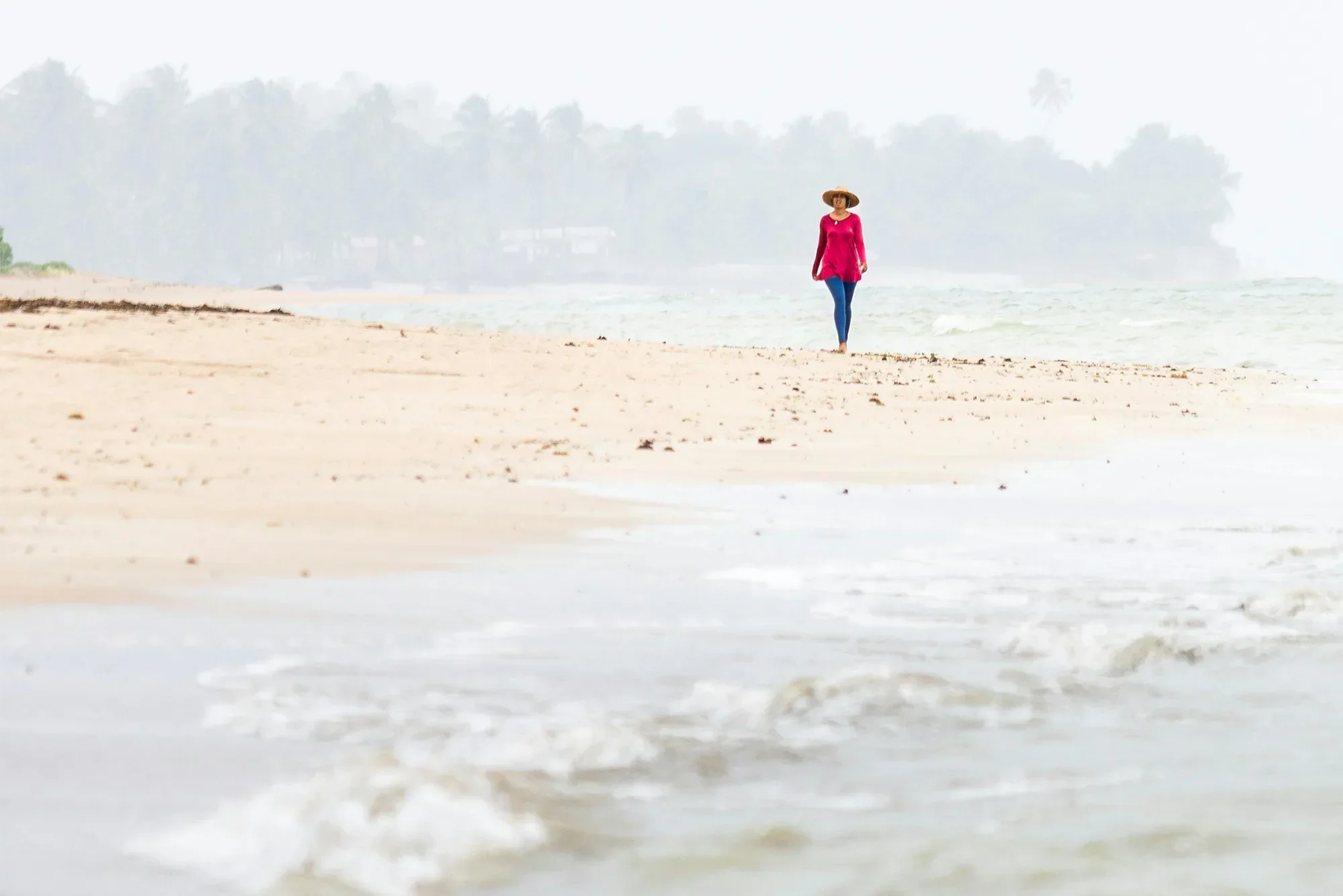 Woman wearing a red jumper, blue knitted pants, and round brim tan colored hat walking alone in the distance on an overcast day at the beach