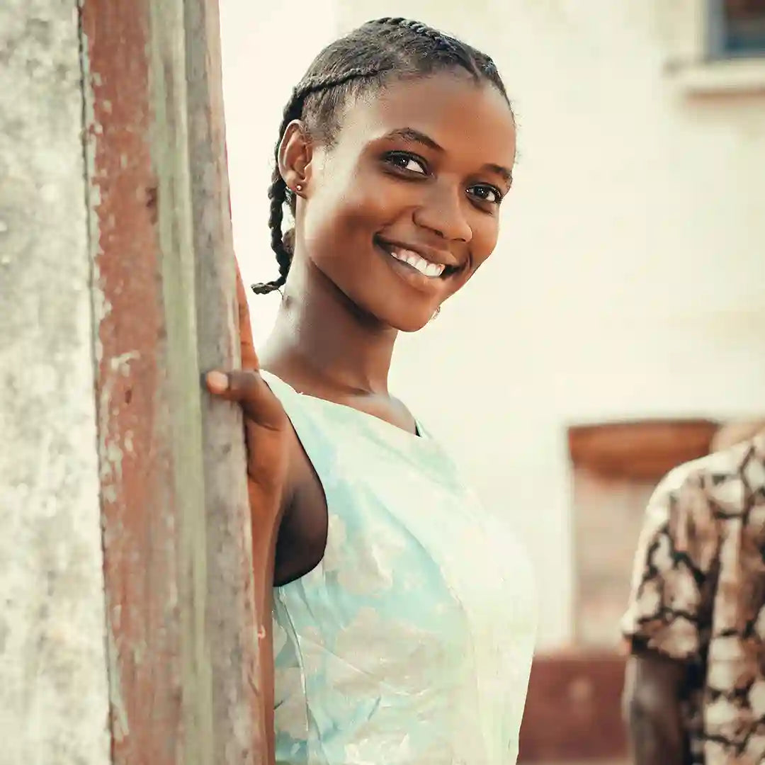 A young African woman with braided hair smiles into the camera while standing outside