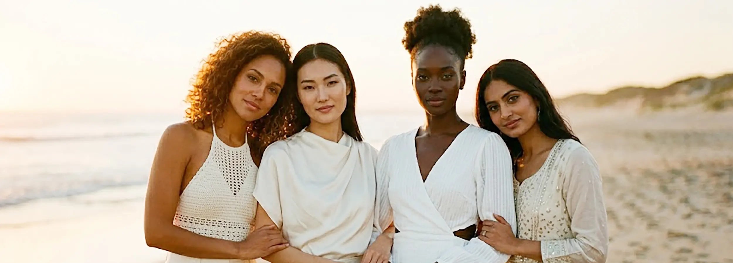 Four multi-ethnic women standing shoulder-to-shoulder in either white or beige colored outfits in the summer during golden hour