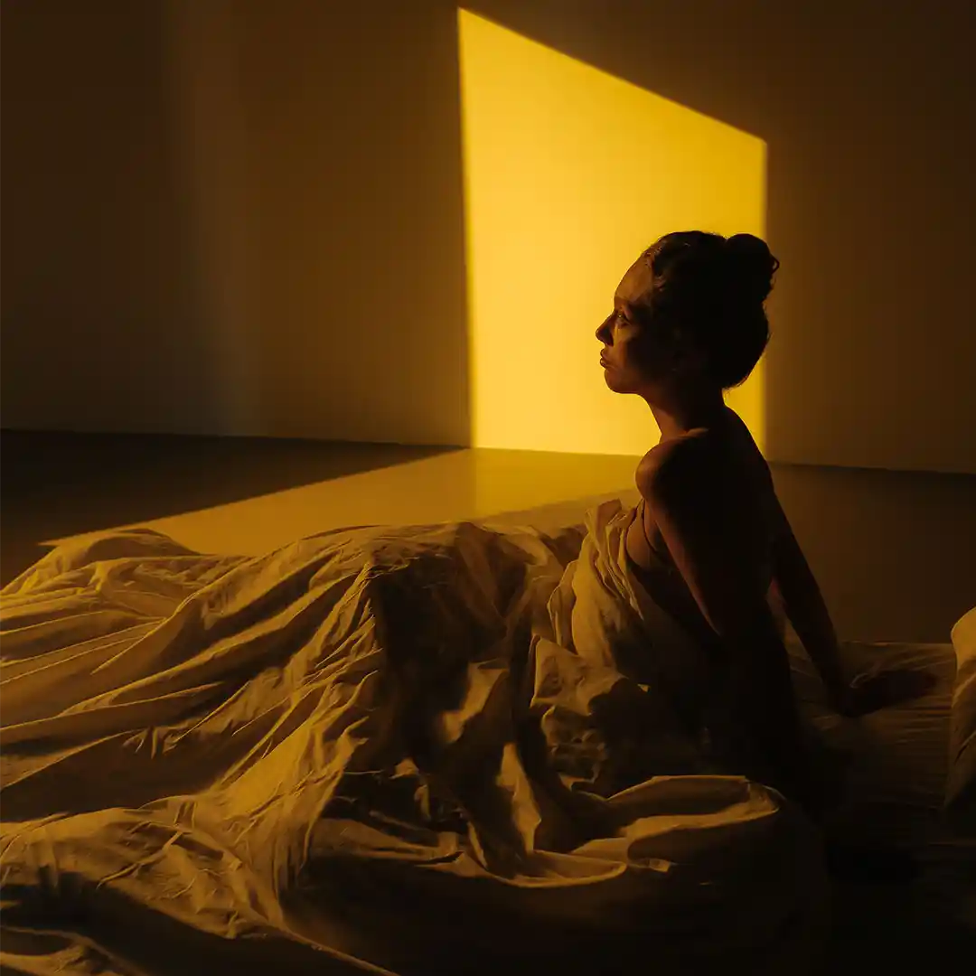 Woman sitting up in bed looking into the yellow light coming from a window