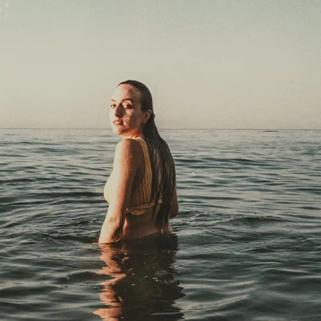 A woman standing alone in the middle of a lake looking over her left shoulder into the camera 