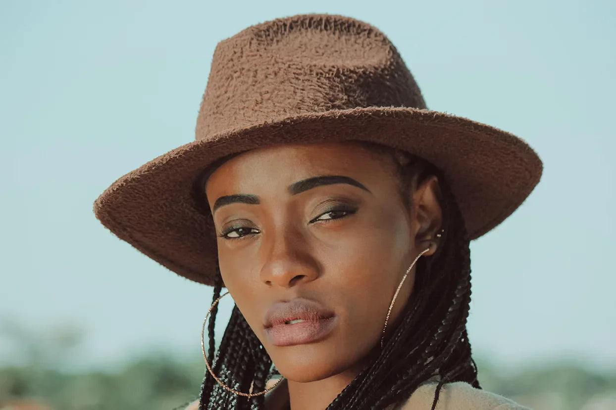 Woman wearing a brown fedora, long braids and large hoop earrings.with large hoop earrings and brown looking directly into camera